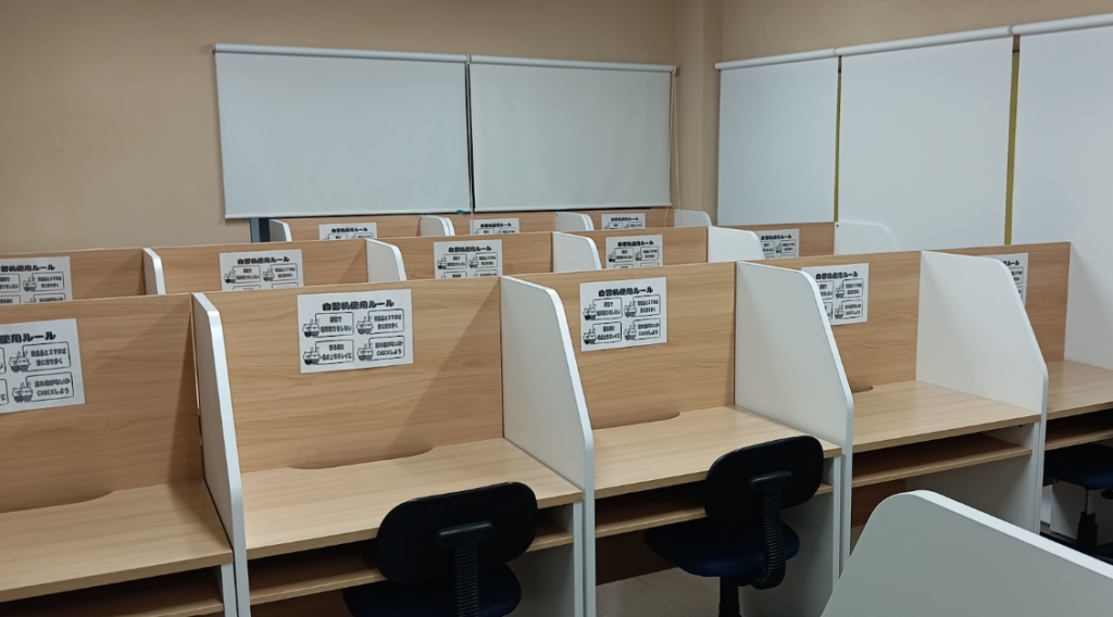 Rows of empty wooden study cubicles with white dividers in a classroom, each cubicle has a posted instruction sign in Japanese and a small black chair.