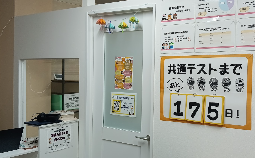 Interior office space with a white door, colorful paper umbrellas hanging above, and a large countdown poster reading '共通テストまで 175 日！' on the wall behind it.