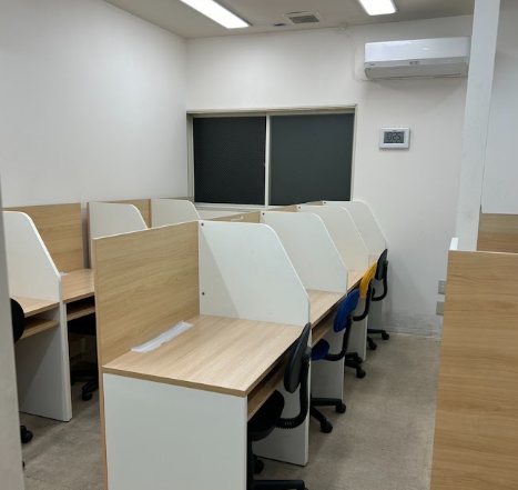 Rows of light wood and white partitioned desks with black and blue chairs in a small office or call-center room.